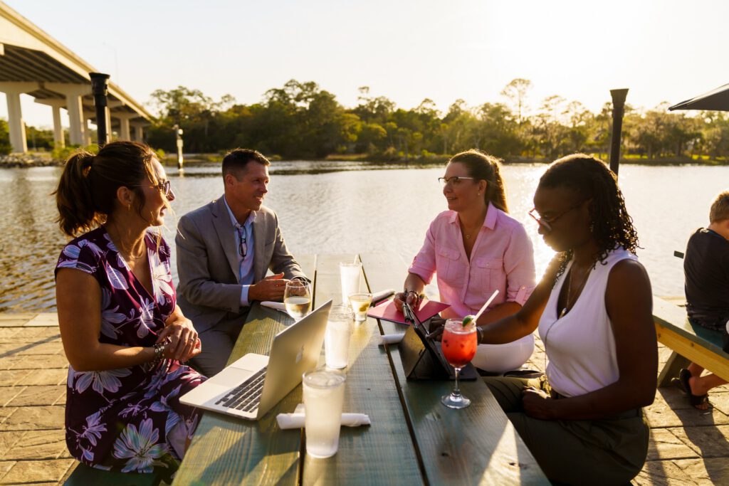Four coworkers share a laugh over a glasses of wine at a shoreside cafe.
