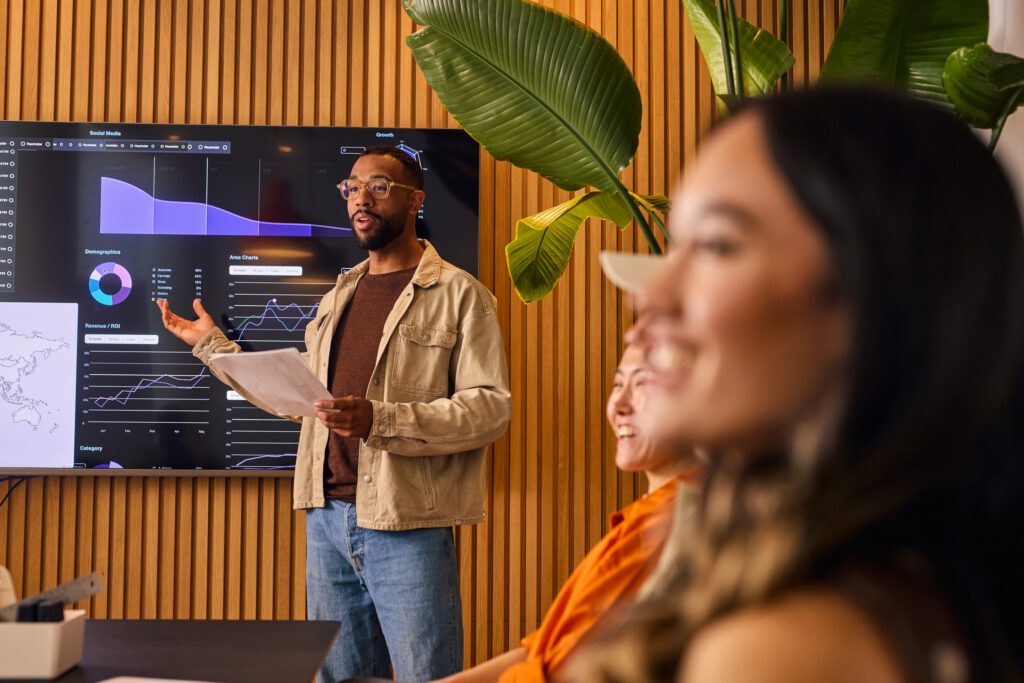 A man presents data on a wall-mounted screen with charts and graphs. He holds papers and gestures to seated colleagues. The setting is modern and professional.