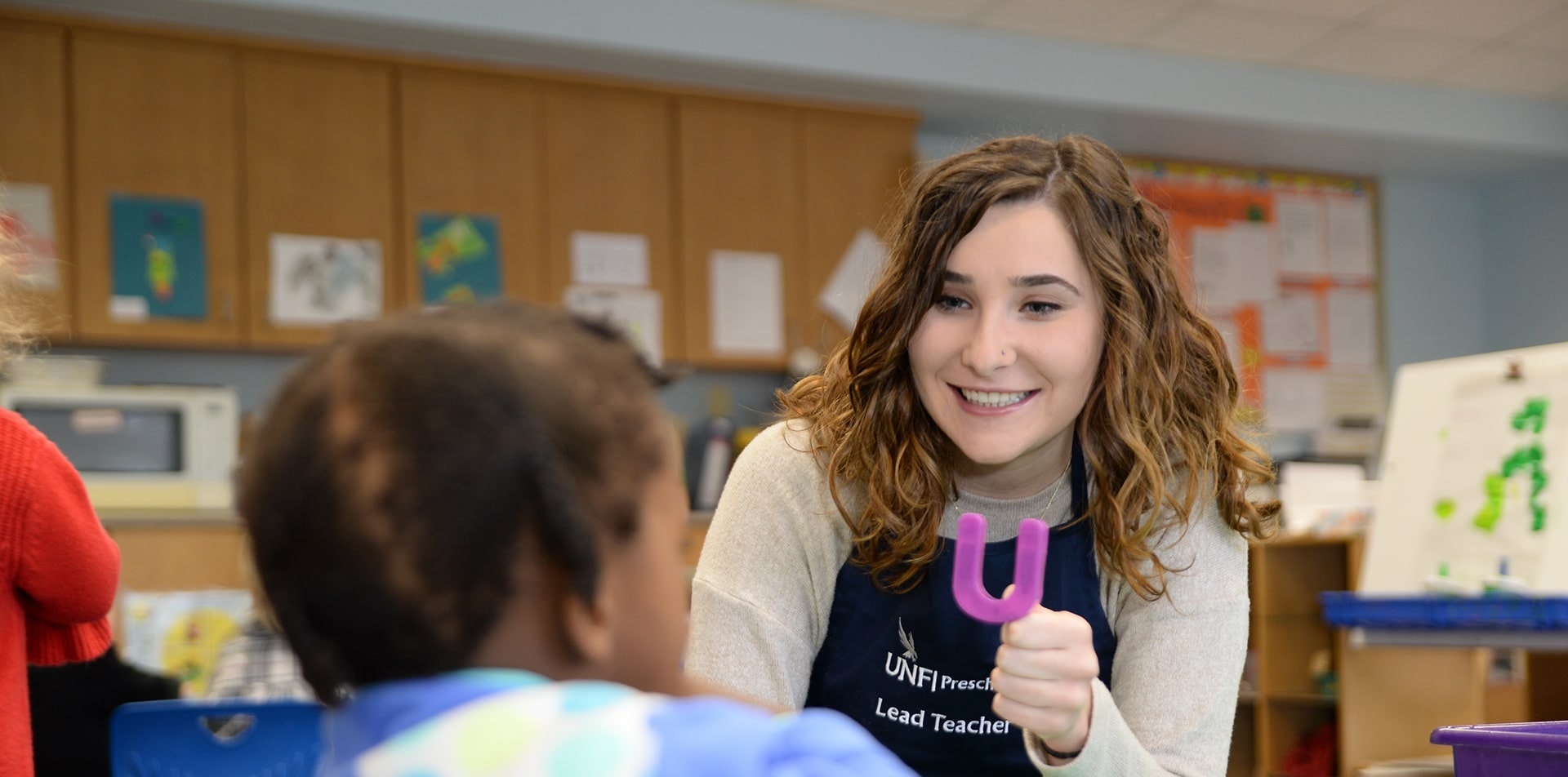 A teacher smiles at a child, holding a purple letter 'U' in a classroom setting. The atmosphere is warm, fostering learning and engagement.