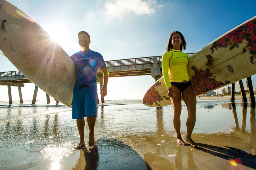 One male and one female surfer emerge from the ocean at the shoreline with sun shining between them.
