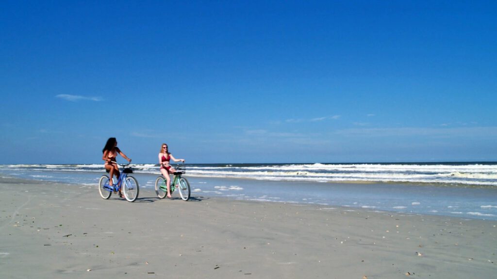 Two women in swimsuits ride bikes along the shoreline.
